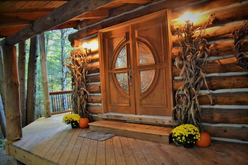 A rustic log cabin entrance with a wooden door, pumpkins, and yellow mums on a wooden porch surrounded by trees.