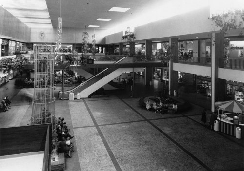 Black and white image of a spacious shopping mall interior with escalators, seating areas, and shops in the background.