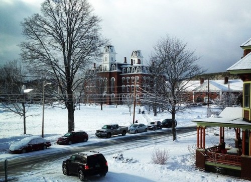 A snowy landscape featuring a historic building, trees, and parked cars along a quiet street.