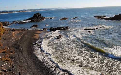 A scenic view of a rocky beach with waves crashing against the shore under a clear blue sky.