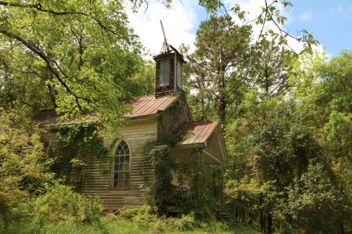 An old, abandoned church overgrown with greenery, featuring a weathered wooden structure and a bell tower.