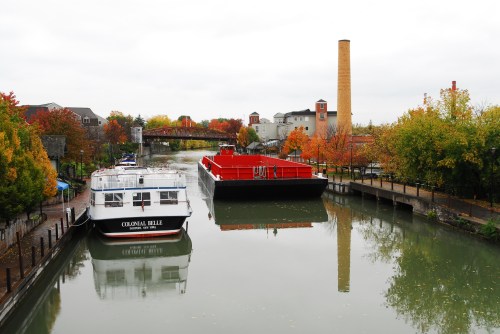 A calm canal scene with colorful autumn trees, a boat named "Colonial Belle," and a tall brick smokestack in the background.