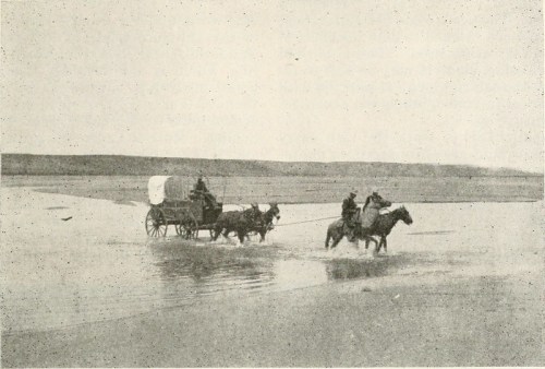 A historical scene of two horse riders guiding a covered wagon through shallow water on a flat landscape.