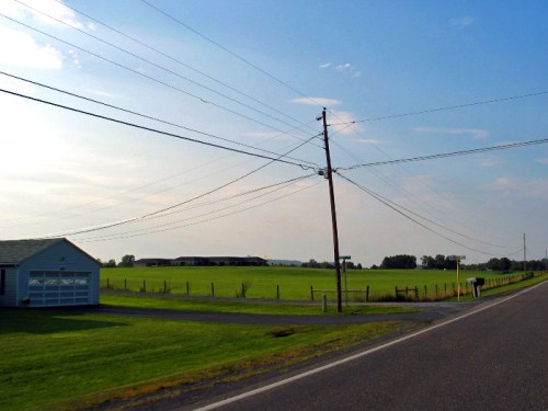 A rural landscape featuring a grassy field, a road, and utility poles under a clear blue sky.