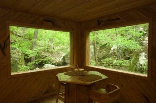 Cozy wooden interior with two large windows showcasing a lush green forest and rocky landscape outside.
