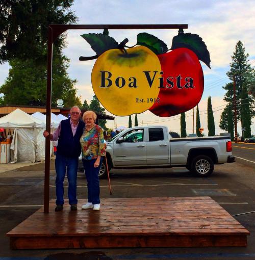 An elderly couple stands beside a colorful sign for Boa Vista, featuring a yellow apple and a red apple.