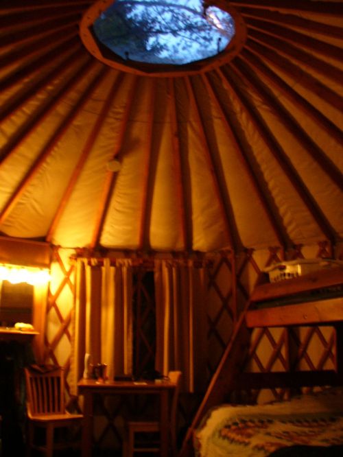 Interior of a yurt with wooden beams, a circular skylight, a table, chairs, and a cozy atmosphere.