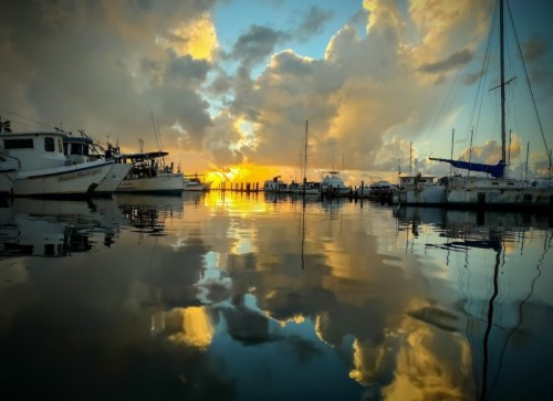Sunset over a calm marina, with boats reflecting on the water and dramatic clouds in the sky.