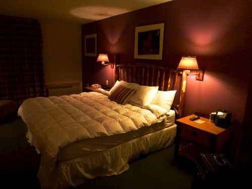 Cozy hotel room with a large bed, warm lighting, and a nightstand, featuring a purple wall and curtains.