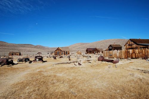 Abandoned ghost town with old buildings and rusted vehicles, set against a clear blue sky and dry, grassy landscape.
