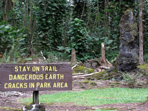 A wooden sign warns to stay on the trail due to dangerous earth cracks, with a forested area and rock formation in the background.