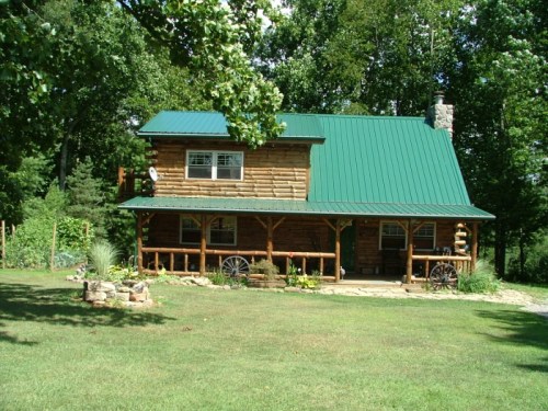 A rustic log cabin with a green metal roof, surrounded by trees and a grassy lawn.