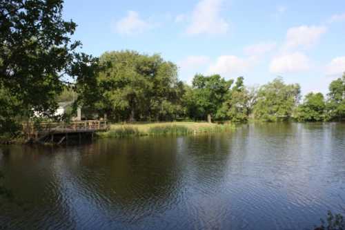 A serene lake surrounded by lush greenery and trees, with a wooden dock on the left under a clear blue sky.