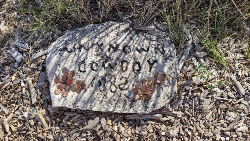 A weathered stone marker reading "Unknown Cowboy 1882," surrounded by scattered pennies and grass.