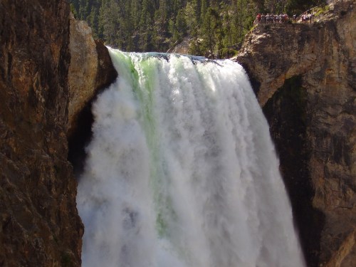 A powerful waterfall cascades down rocky cliffs, surrounded by lush green trees and a crowd of visitors above.