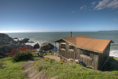 A rustic wooden cabin overlooks a rocky coastline and the ocean, surrounded by green grass and vibrant red flowers.