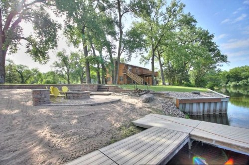 A serene riverside scene featuring a wooden house, sandy area, yellow chairs, and a dock surrounded by lush greenery.