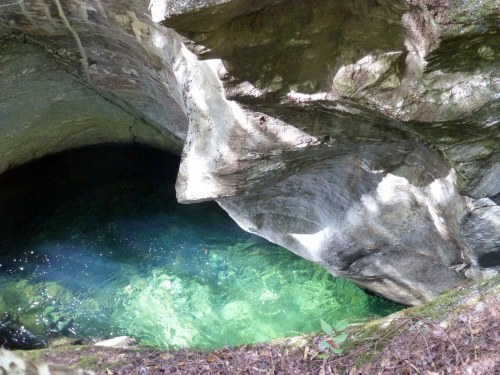 A clear, greenish pool of water surrounded by rocky cliffs and natural vegetation. Sunlight reflects on the water's surface.