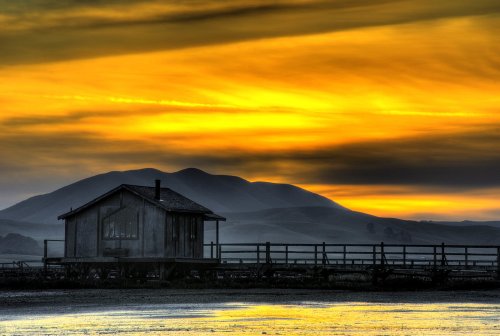 A rustic cabin by the water at sunset, with vibrant orange and yellow skies and distant hills in the background.