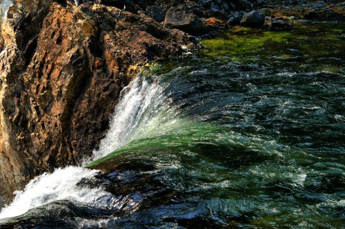 A close-up of a rocky riverbank with water cascading over rocks, creating ripples in a clear, greenish stream.