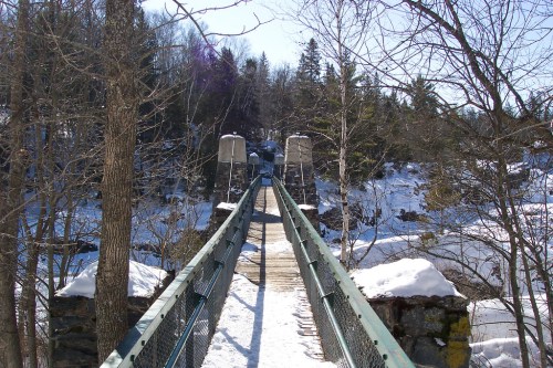 A snowy bridge stretches over a river, surrounded by trees and rocky terrain under a clear blue sky.