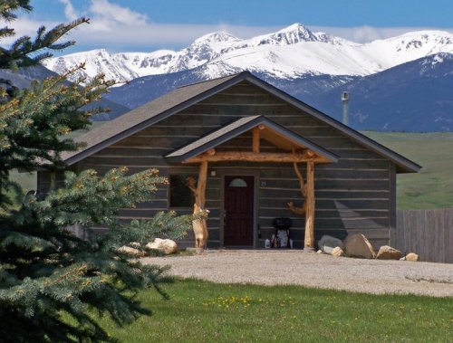 A rustic wooden house with a log porch, set against a backdrop of snow-capped mountains and a clear blue sky.