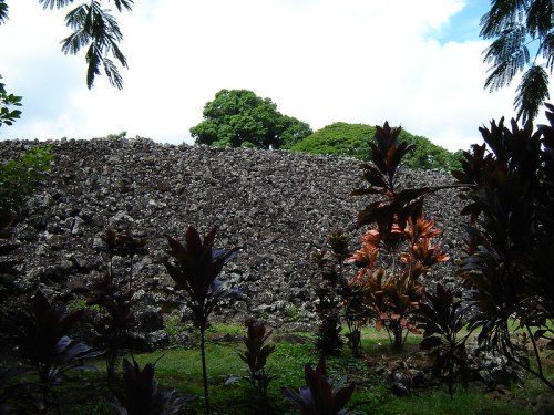 Ancient stone structure surrounded by lush greenery and trees under a partly cloudy sky.