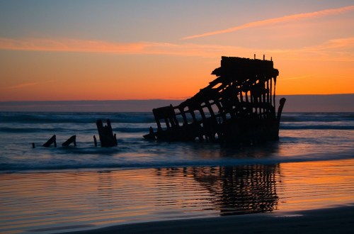 Silhouette of a shipwreck on a beach at sunset, reflecting in calm waters with colorful sky.