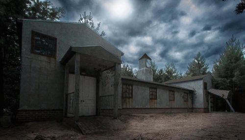 An eerie, abandoned building surrounded by trees under a cloudy sky, with a sandy foreground.