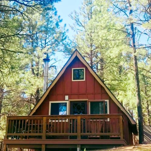 A cozy red cabin with a wooden deck, surrounded by tall green trees under a clear blue sky.