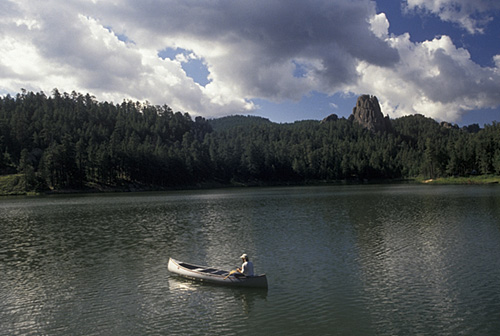 A person paddles a canoe on a calm lake surrounded by lush forests and rocky hills under a partly cloudy sky.