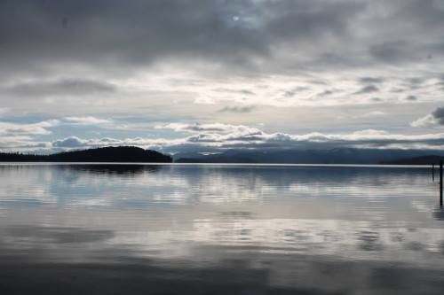 A serene lake scene with calm waters reflecting cloudy skies and distant mountains under a grayish atmosphere.