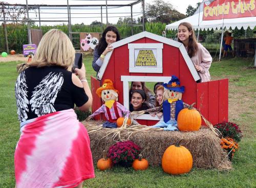 A group of girls poses in a barn cutout with scarecrows and pumpkins, while a woman takes their photo.