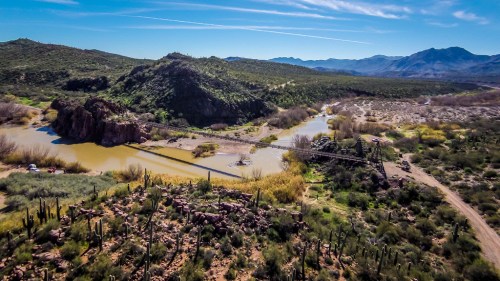 Aerial view of a river winding through a desert landscape with mountains, greenery, and a bridge in the distance.