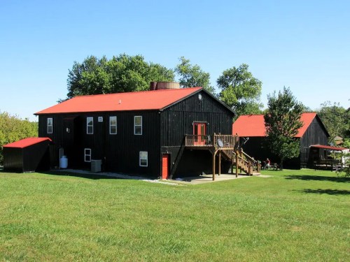A black wooden house with a red roof, surrounded by green grass and trees under a clear blue sky.