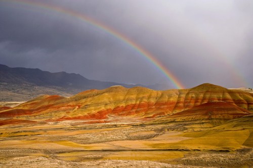 Colorful striped hills under a cloudy sky, with a vibrant rainbow arching above the landscape.