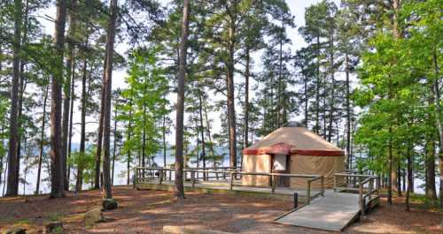 A yurt surrounded by tall trees, with a wooden deck overlooking a serene lake.