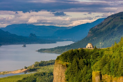 A scenic view of a river winding through mountains, with a historic building perched on a cliff surrounded by lush greenery.