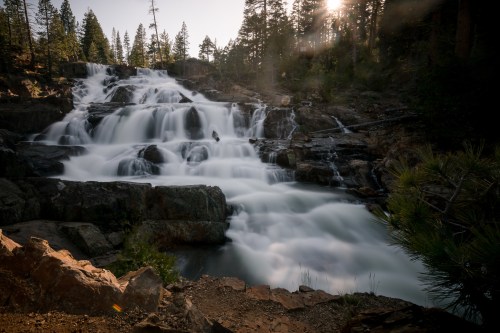 A serene waterfall cascades over rocks, surrounded by trees and bathed in soft sunlight.