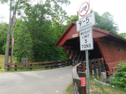 A red covered bridge with a clearance sign and weight limit sign, surrounded by greenery and a gravel road.
