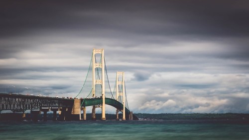 A bridge spans a body of water under a cloudy sky, with green accents and tall towers.