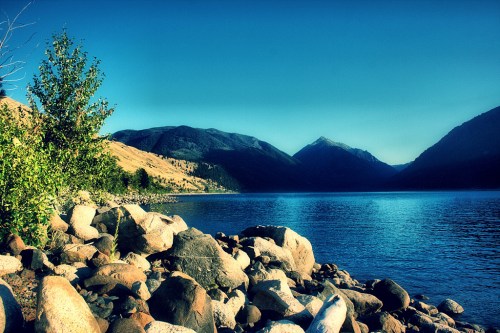 A serene lake surrounded by mountains, with rocky shores and lush greenery under a clear blue sky.