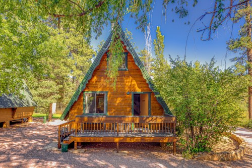 A charming A-frame cabin surrounded by trees and greenery, with a wooden porch and clear blue sky.
