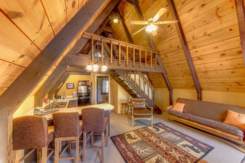 Cozy A-frame cabin interior with wooden walls, a loft, bar seating, and a living area featuring a rug and sofa.