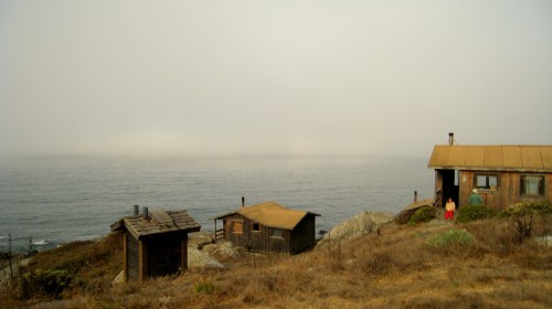 A foggy coastal scene with two wooden cabins overlooking the ocean and a person in red shorts near the water's edge.
