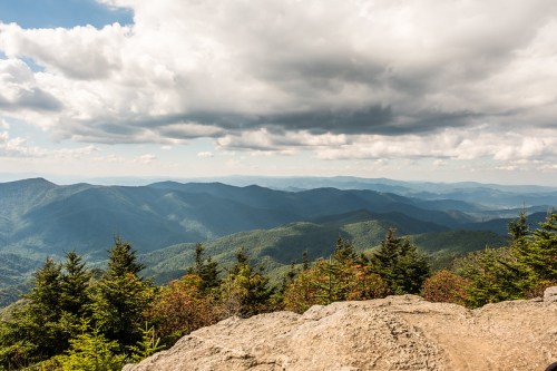A panoramic view of rolling mountains under a cloudy sky, with lush greenery and rocky foreground.