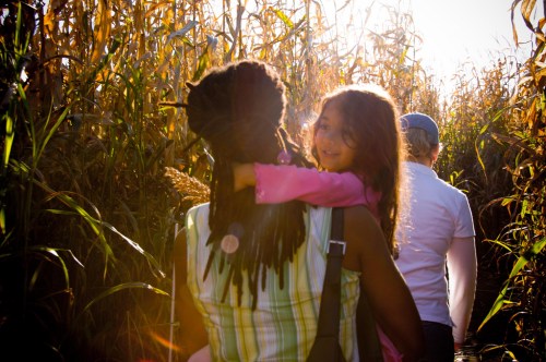 A woman carries a girl on her back, walking through a sunlit cornfield with another person in the background.