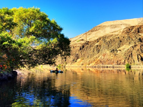 A serene river scene with a kayaker, surrounded by rocky hills and lush greenery under a clear blue sky.