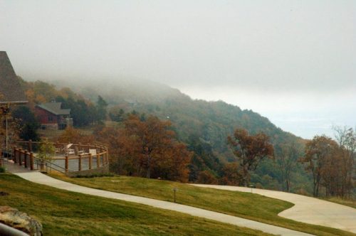 Foggy landscape with autumn trees, a winding path, and a distant view of hills and a cloudy sky.