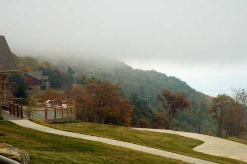 A foggy landscape with autumn trees, a winding path, and a distant view of hills and a house.